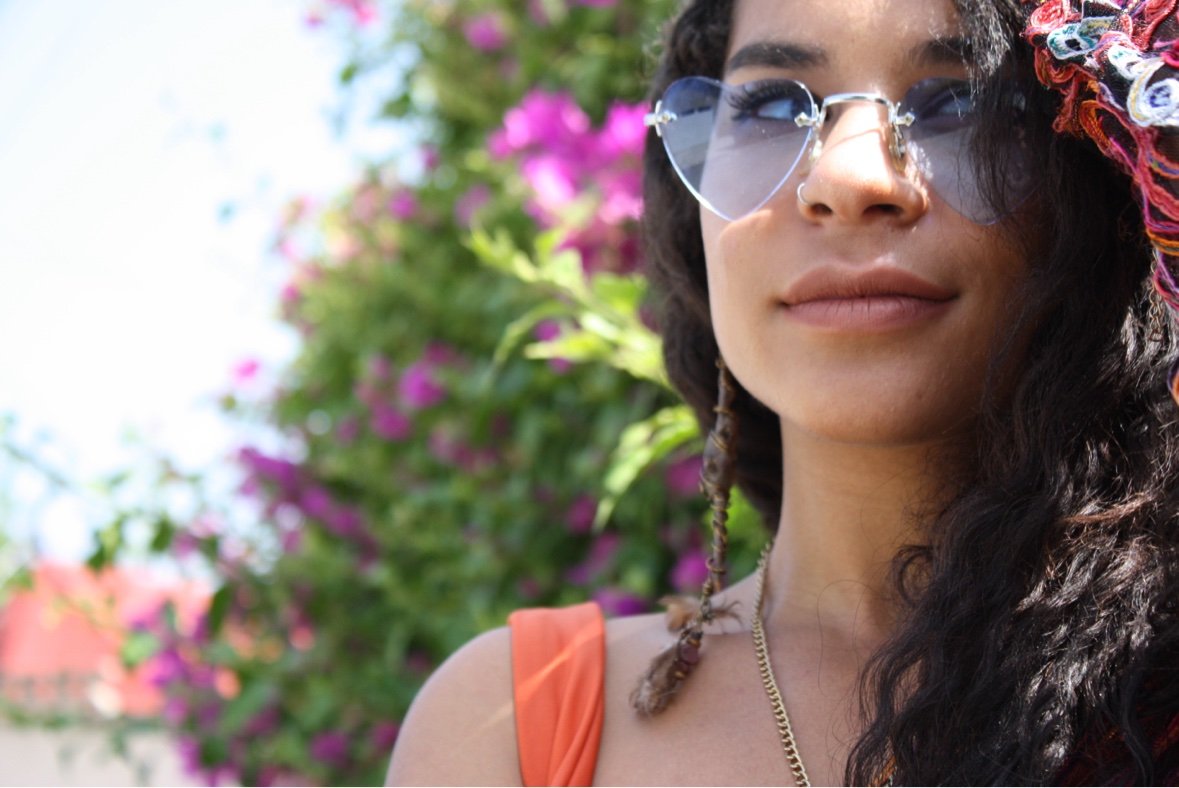 A woman in heart-shaped sunglasses and a gold chain necklace, with magenta bougainvillea behind her