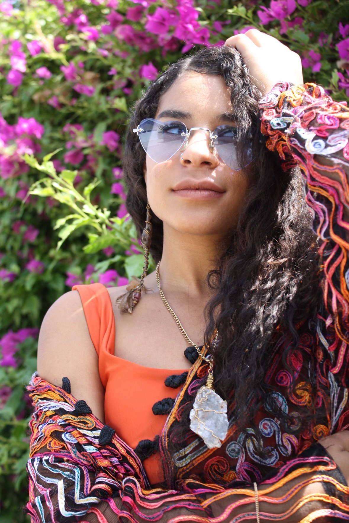 A woman in heart-shaped sunglasses wearing a wire-wrapped quartz pendant, with bougainvillea behind her