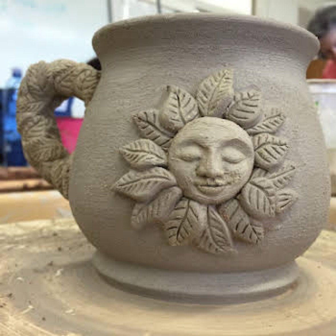 An unfired clay mug with a sculpted sun face and leaf-textured handle