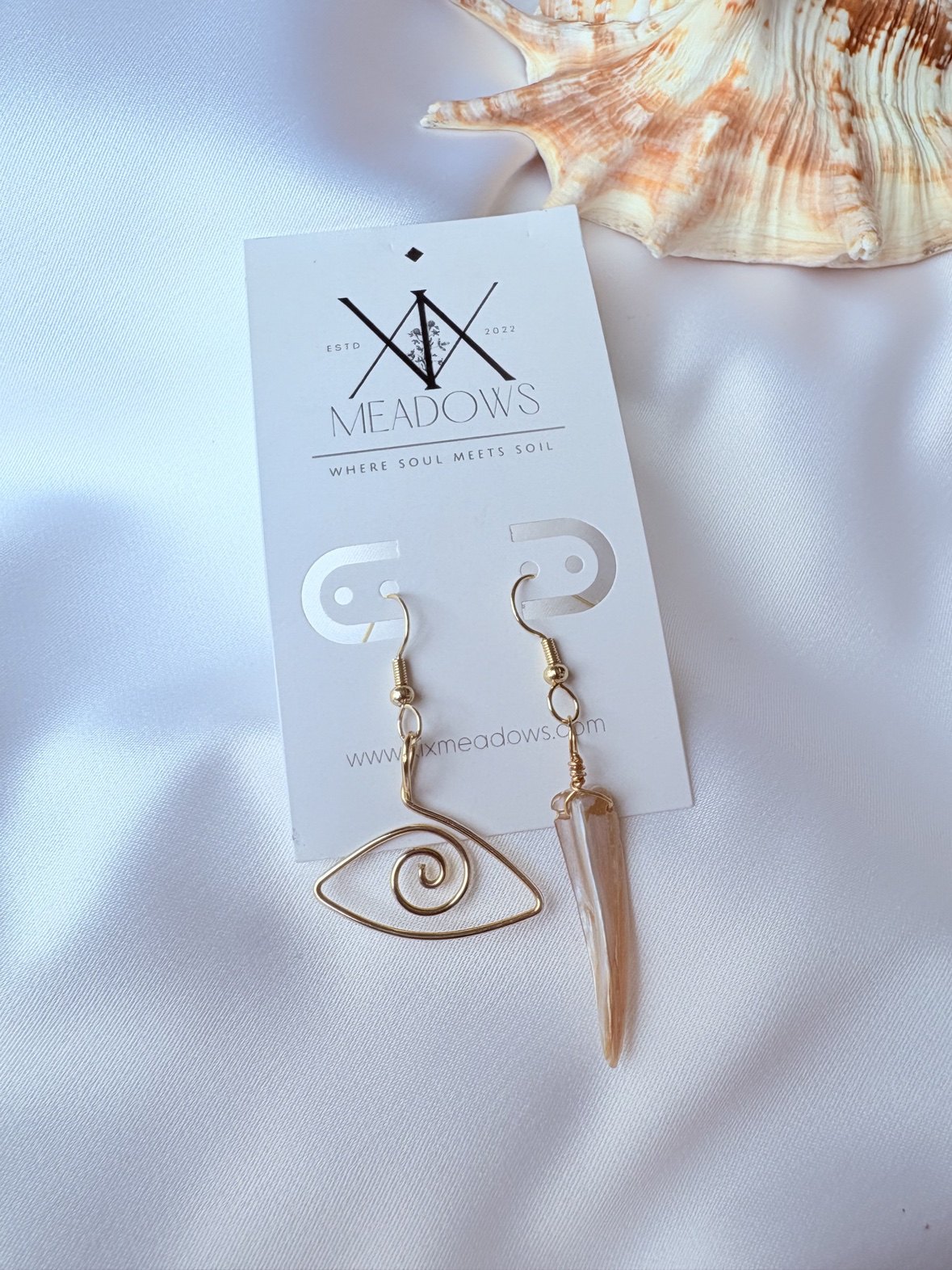 Mismatched gold earrings — a spiral eye and a shell spike — on a XiX Meadows card