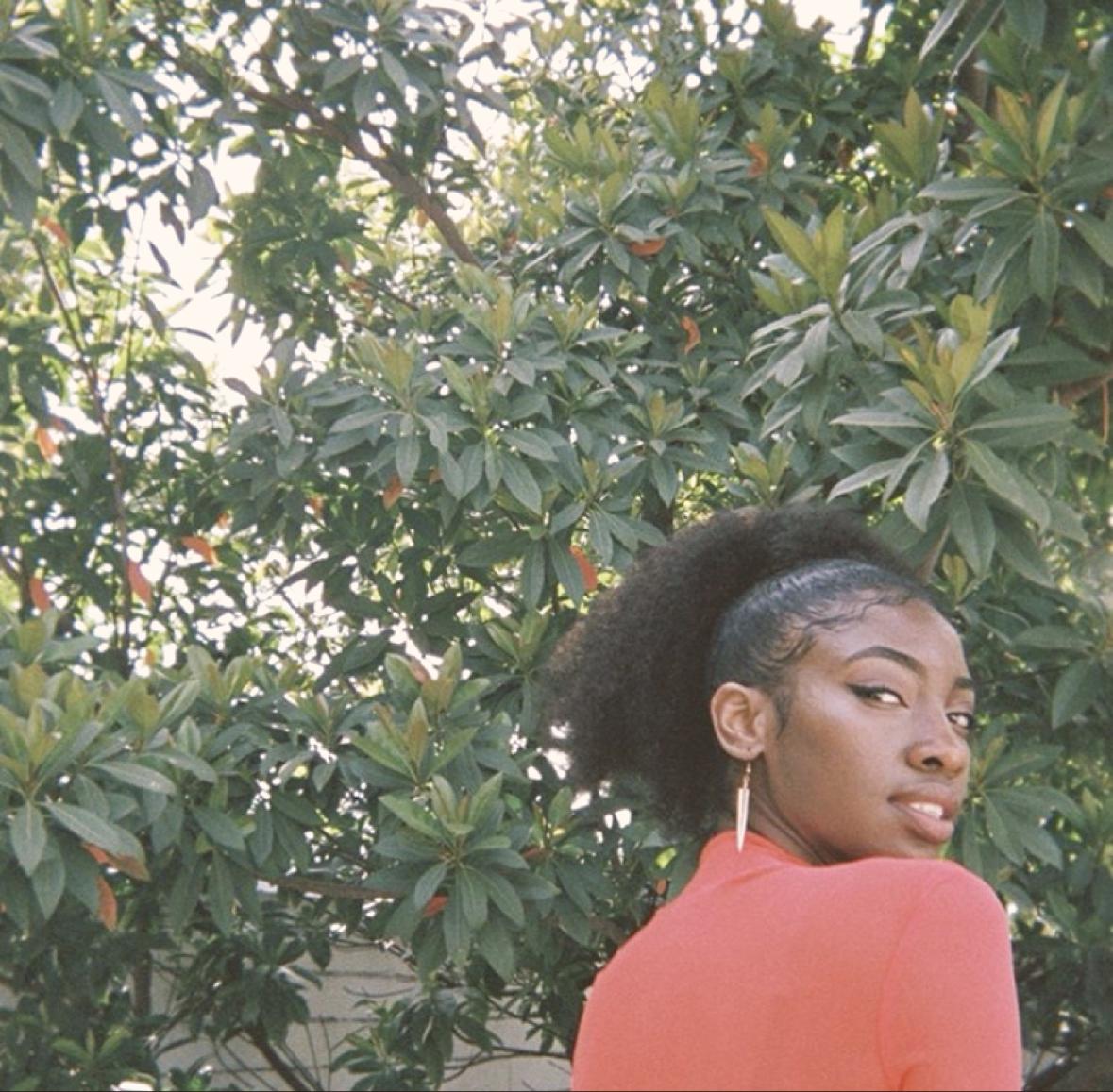 A person in a red top with greenery behind, wearing a long copper leaf earring